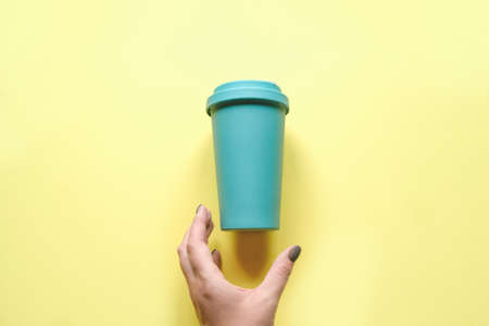 A woman's hand reaches for a mint-colored coffee mug. Bamboo glass on yellow background. Recyclable material, zero wasteの写真素材