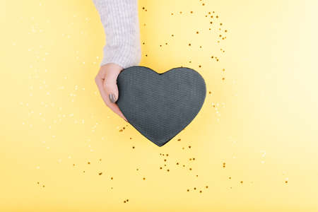 A woman's hand holds a gift in the form of a black heart on a beautiful yellow background with sequinsの写真素材