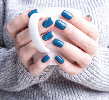 Mug of coffee or tea in the hands of a woman dressed in a knitted sweater. Beautiful women's manicure blueの写真素材