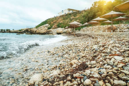 Pebble beach and beautiful waves with white foam. Beach umbrellas made of straw, the rocky coast of the island of Crete in the setting sun. Fantastic seascape landscapeの写真素材