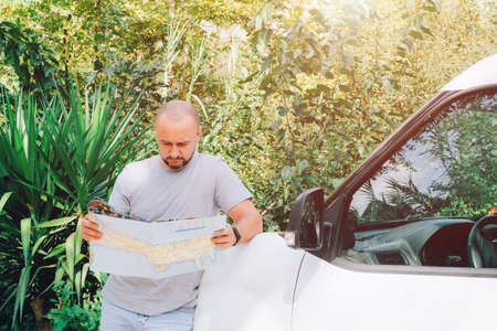 A young man stands near the car with a map of the area in his hands. Travel by car. Planning your vacation and route. Beautiful background of tropical greenery. Car for rentの写真素材
