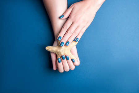 Well-groomed female hands on a blue background. Neat blue nail Polish. Starfish in hand. Manicure for a trip to the sea. Stylish manicure, great design for any purposes. の写真素材