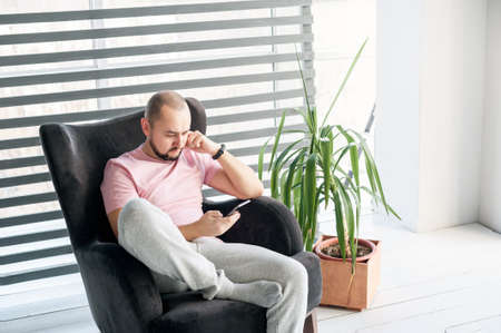 Man sits in armchair and looks at the phone. His view is serious and focused. The pose is relaxed. Home cloth: T-shirt, sweatpants, socks. Background is window with shutters, plant in flowerpot.の写真素材