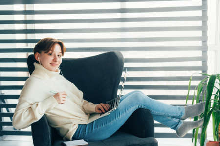Teen girl sitting in a chair with a laptop in headphones and looking at the screen. Video Conversationの写真素材