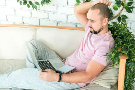 A man in home clothes is sitting with a laptop on the couch. The concept of remote work and educationの写真素材
