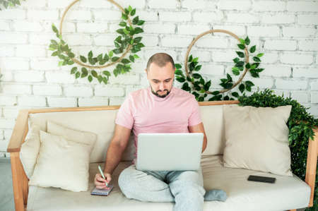 A young man with a beard is sitting on the sofa, looking at the laptop screen and making notes in a notebookの写真素材