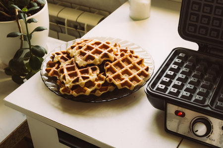 Stack of golden waffles on glass plate next to waffle maker for breakfast menu, recipe, culinary blog and food background.の写真素材