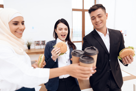 A woman in a hijab with her colleagues eating a hamburger and drinking coffee. They are in a good mood. They have a break at work.の写真素材