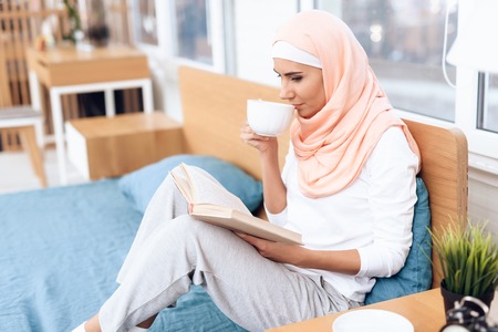 An arabian woman is drinking tea and reading a book while sitting on the bed. She rests at home.