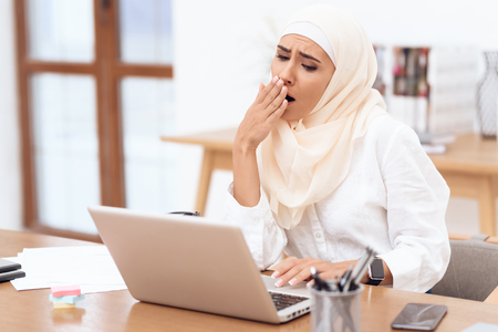 Arab woman wearing a headscarf sitting tired at work. She yawns. She is sitting at the desk in the office.の写真素材