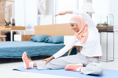 Arab woman doing gymnastics in the bedroom. She's at home. She has a headscarf on her head.