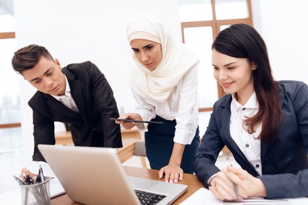 The Arab woman in hijab works in the office together with her colleagues. They are engaged in business. On the desk is a laptop.の写真素材