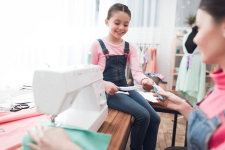 A woman sews on a sewing machine. Next to her is a little girl. They are with mom in the sewing workshop.の写真素材