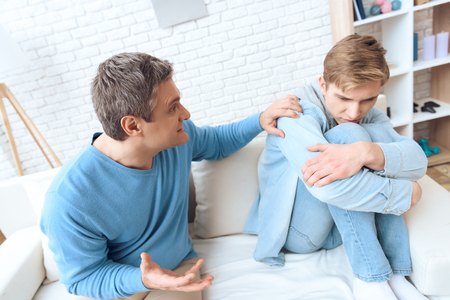 Father sitting near on couch, tries to talk to his son, but problem teenager doesn't want to listen. Father reaches out with his hand.の写真素材