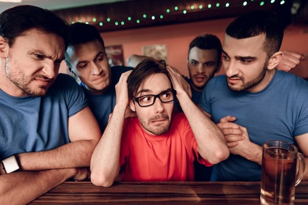 Blue team sports fans at bar surrounding red team fan at sports bar. They are watching football game.の写真素材