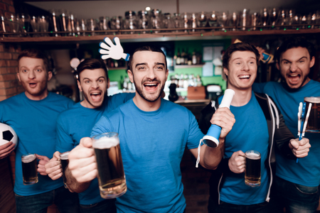 Five sports fans drinking beer celebrating and cheering at sports bar. They are supporting blue team.の写真素材