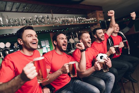 Soccer fans sitting in line celebrating and cheering drinking beer at sports bar. They are supporting red team.の写真素材