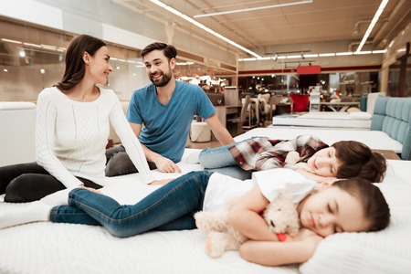 Young lovely couple is sitting on mattress next to sleeping children in furniture store. Testing softness of mattress. Choosing mattress in store