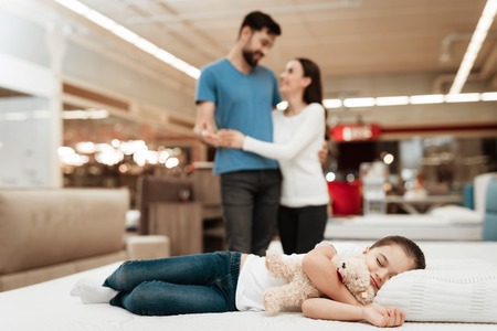 Young happy couple in background enjoying sleeping little girl. Choosing mattress in store. Peaceful little girl sleeps on mattress and hugs teddy bear.の写真素材