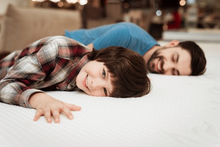 Handsome bearded father with young son is testing mattress for softness. Choosing softness of mattress. Choosing mattress in storeの写真素材