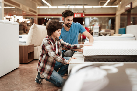 Bearded man with young son is testing mattress for softness. Testing softness of mattress. Choosing mattress in storeの写真素材