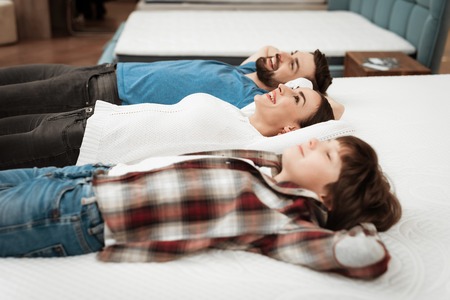 Young happy couple with cute little boy lies on bed in mattress store. Family checking softness of mattress. Purchase of orthopedic mattress.の写真素材