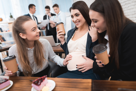 Three young beautiful girls communicate on coffee break and eat sweet cakes. Young pregnant woman with girlfriends drinking latte at lunch break.の写真素材