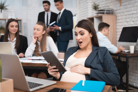 Surprised pregnant girl looks at tablet in office. Pregnancy at work. Pregnant woman at her desk. Pregnant woman in good mood. Brainstorm at work.の写真素材