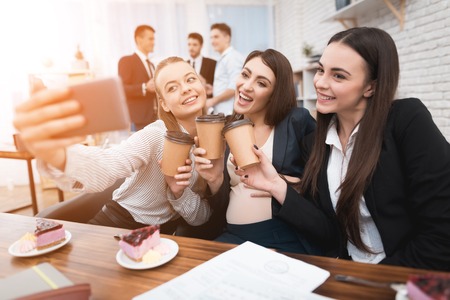 Two young beautiful girls together with pregnant woman doing selfie on phone. Pregnant woman does selfie at office. Three beautiful girls are posing in front of camera phone.の写真素材