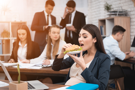Beautiful pregnant girl eats sandwich. Adult pregnant girl caress pregnant belly. Pregnant woman in office. Pregnancy in work.の写真素材