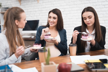 Three young cute girls eating sweet cake at lunchtime in office. Lunch break. Coffee break in office. Pregnant woman with young girlfriends dines sweet.の写真素材