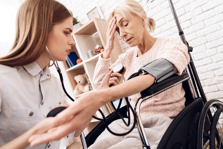 Girl is caring for elderly woman in wheelchair at home. Girl is checking blood pressure with tonometer.の写真素材