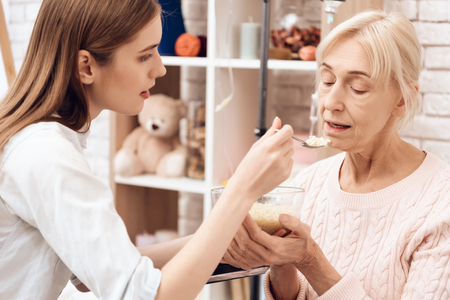 Girl is nursing elderly woman in wheelchair at home. Girl is helping woman with dinner.の写真素材