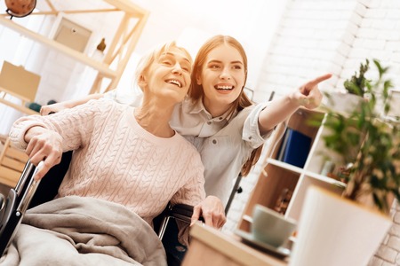 Girl is nursing elderly woman at home. Girl is riding woman in wheelchair in living room. They are happy.の写真素材