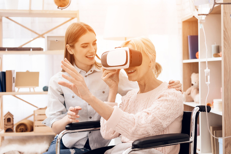 Girl is caring for elderly woman in wheelchair at home. Woman is using virtual reality.の写真素材
