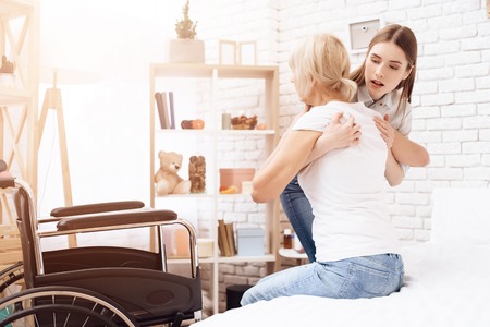 Girl is caring for elderly woman at home. Girl is helping woman to get from bed into wheelchair.の写真素材