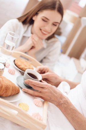 Girl is caring for elderly woman in bed at home. Girl brings breakfast on tray. Woman is drinking coffee.の写真素材