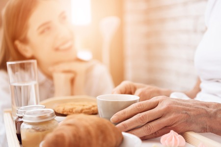 Girl is caring for elderly woman in bed at home. Girl brings breakfast on tray. Girl is smiling.の写真素材