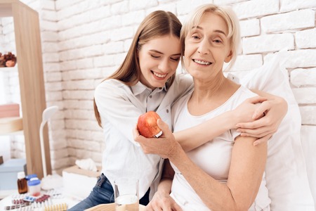 Girl is nursing elderly woman in bed at home. They are embracing. Woman is holding apple.