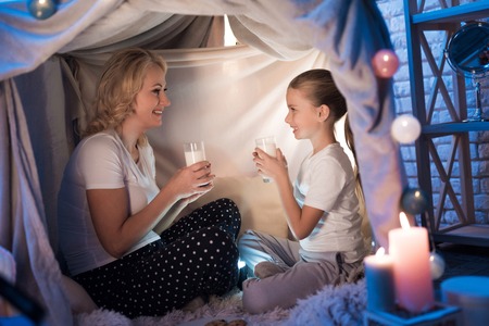 Grandmother and granddaughter are eating cookies with milk in cozy blanket house at night at home.の写真素材