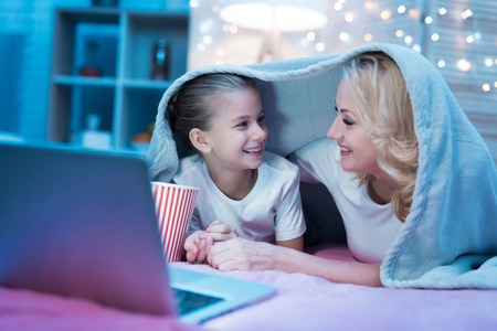 Grandmother and granddaughter are watching movie on laptop under blanket on bed at night at home.の写真素材
