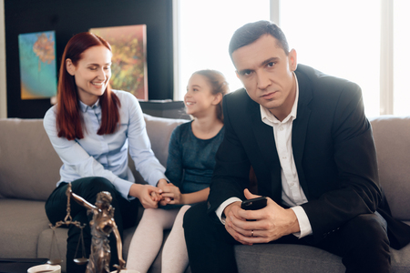 Frustrated father with phone in hands sits on couch next to young wife and daughter. Dissolution of marriage of two adults. Divorce of adult couple.の写真素材