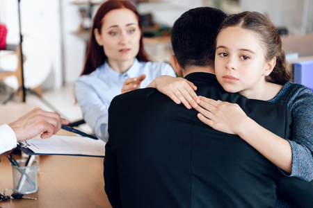 Little girl hugs man in suit sitting opposite woman in lawyer's office. Family in office of family lawyer. Young parents argue about one who will take guardianship of daughter in lawyer's office.の写真素材