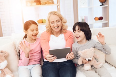 Joyful young grandmother with grandchildren in mood sends greetings to someone on tablet. Grandmother with grandchildren communicates via videoconference on tablet.の写真素材
