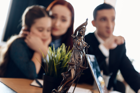 Bronze statue of Themis stands on table, behind which sits divorcing parents with little girl. Young man and beautiful woman decide who will be main guardian of little girl.の写真素材