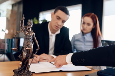 Statue of Themis holds scales of justice. In unfocused background, adult man signs documents. Divorcing couple dissolves marriage contract. Couple going through divorce signing papers.の写真素材