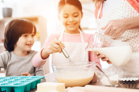Happy boy looks at little girl who is beating eggs in bowl, where beautiful grandmother pours milk from jug. Grandmother teaches grandchildren to cook.の写真素材