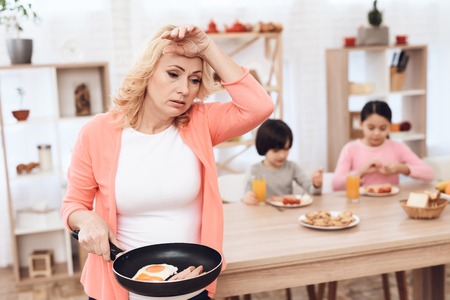 Tred old woman holds frying pan with fried sausages and eggs, standing in kitchen with children. Grandmother prepared dinner for her grandchildren.の写真素材