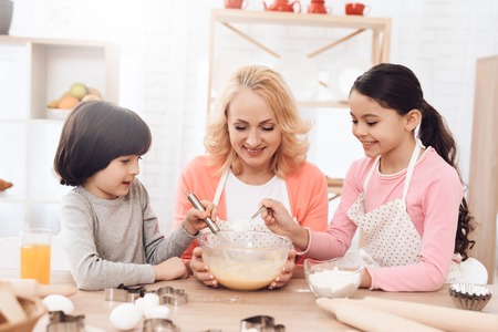 Joyful little boy is beating eggs in bowl with milk and her sister is pouring flour. Grandmother teaches grandchildren to cook cookies in kitchen. Children add ingredients to bowl to make cookies.の写真素材