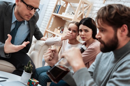 Anxious mother and daughter look at father of alcoholic sitting at reception with psychologist. Discussion of family problems in office of psychologist.の写真素材